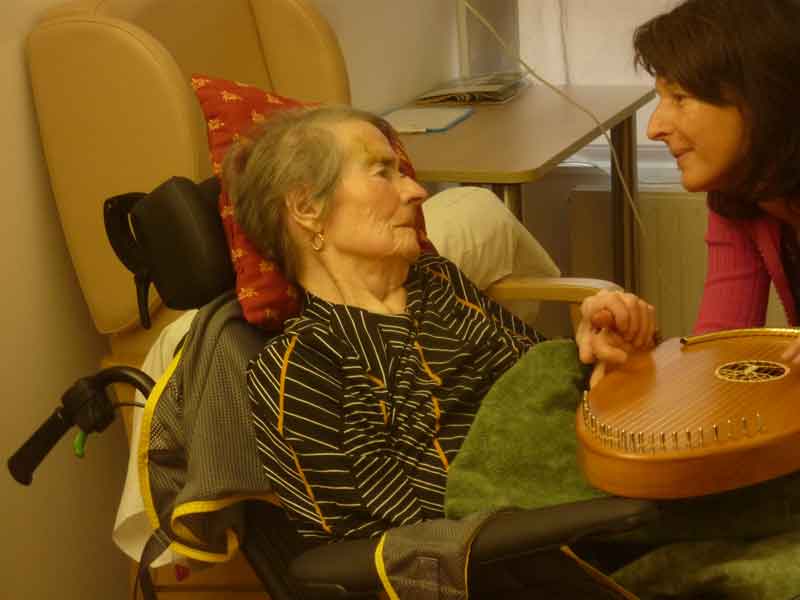 Une femme regarde avec affection une personne âgée assise dans un fauteuil roulant, un instrument de musique entre elles.